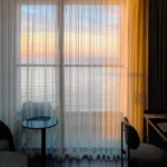 A hotel room with golden sunset light glowing through sheer curtains and a view of the sky