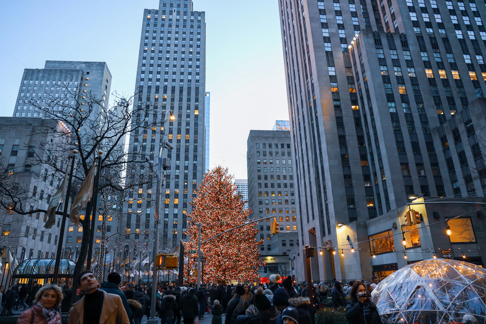 Pedestrians on a Midtown street with the lit Rockefeller Center Christmas tree in the distance at dusk