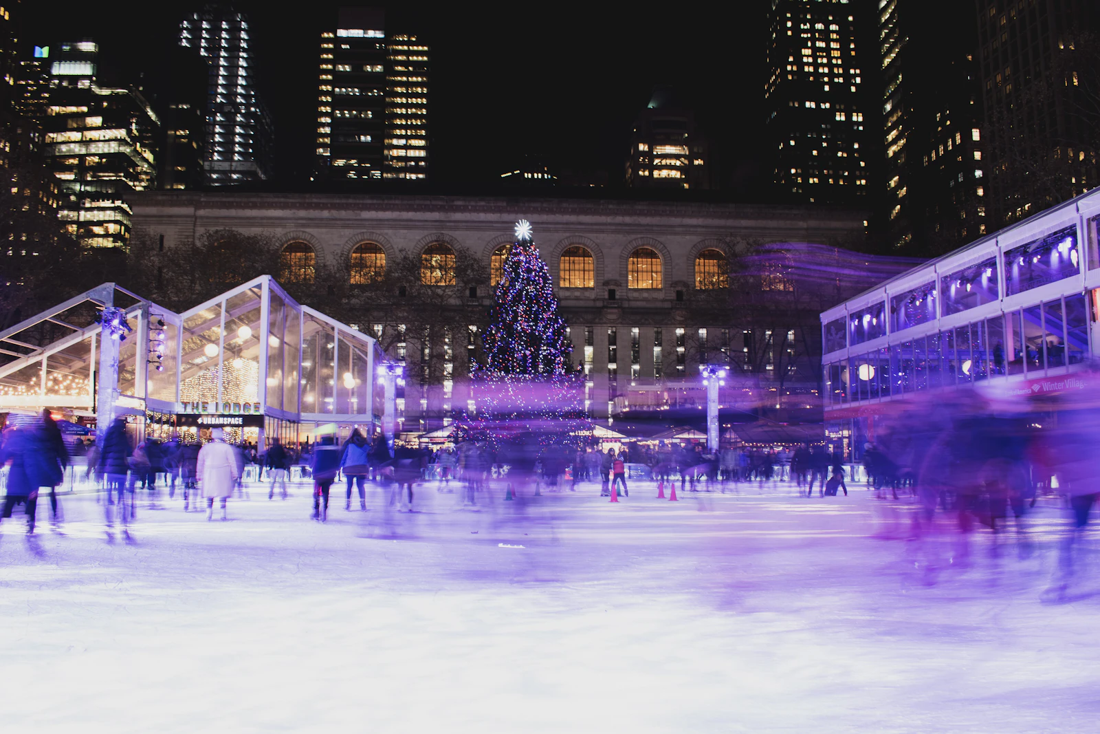 Blurred skaters on the Bryant Park ice rink at night with a lit tree and the library behind