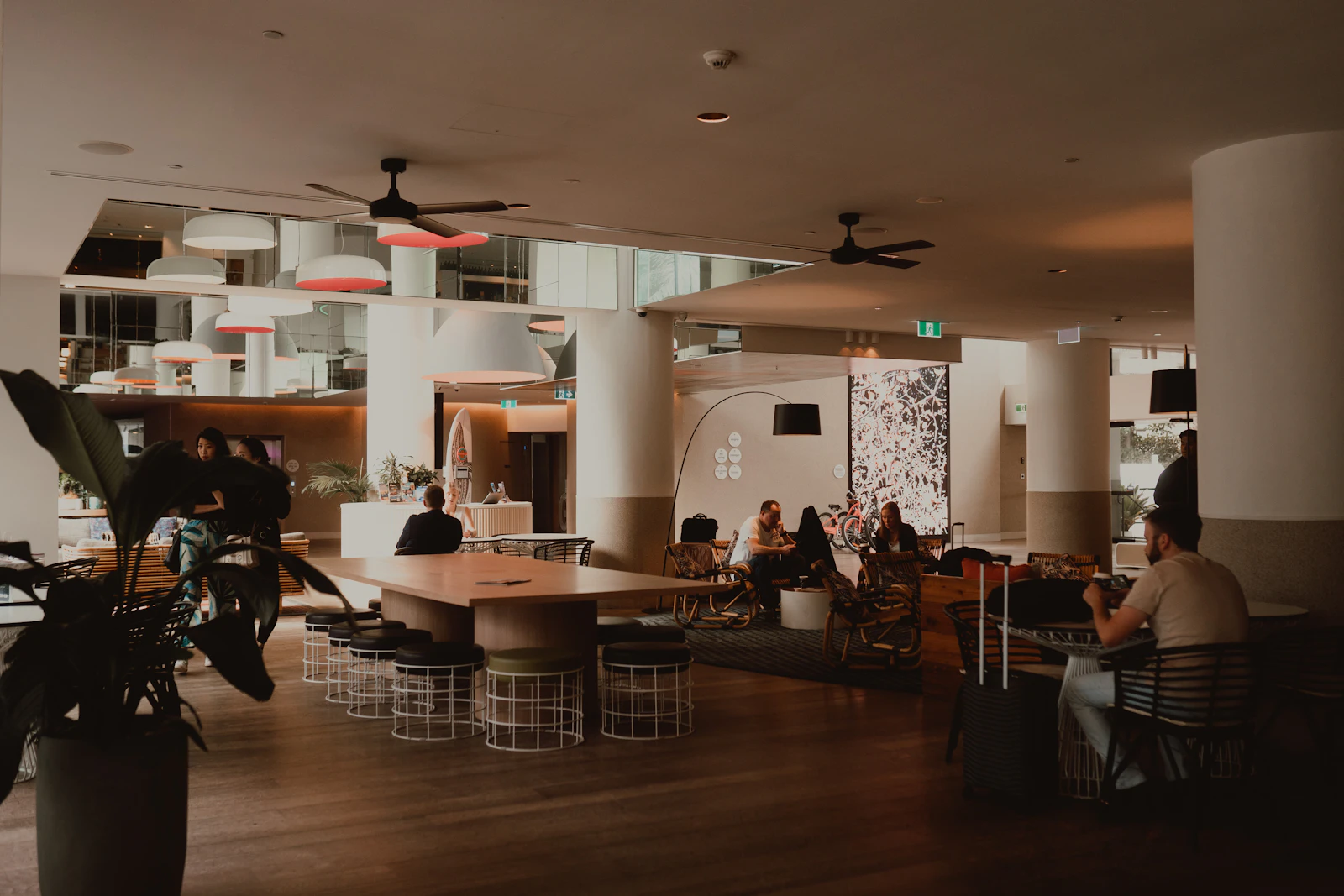 People sitting at tables in a spacious hotel lobby lounge with a long wooden table and ceiling fans