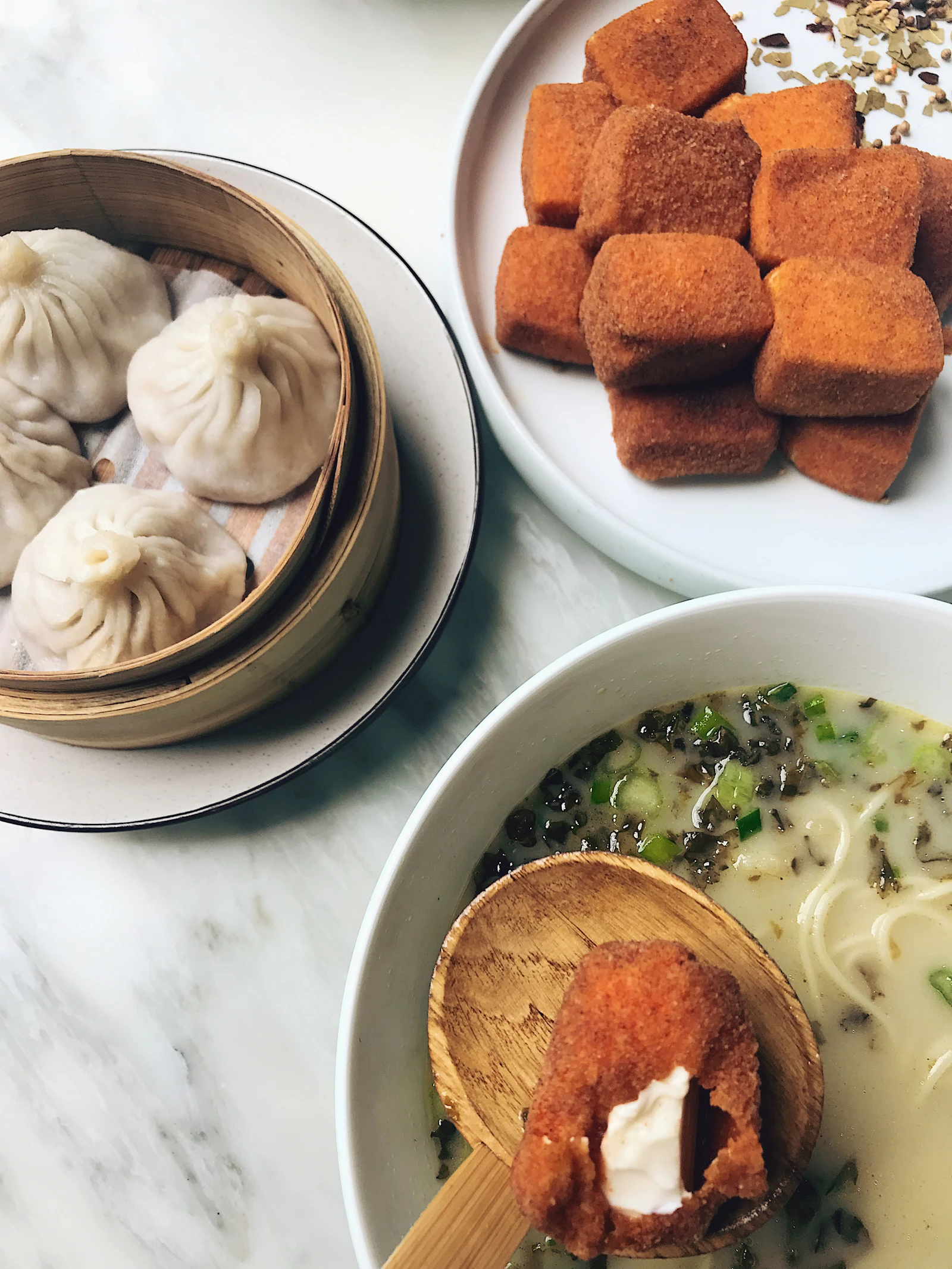 Soup dumplings in a bamboo steamer beside a bowl of noodle soup on a marble table