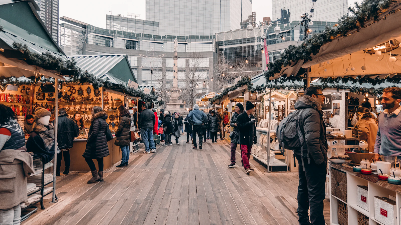 Shoppers walking between wooden holiday market stalls at Columbus Circle with the monument and buildings behind