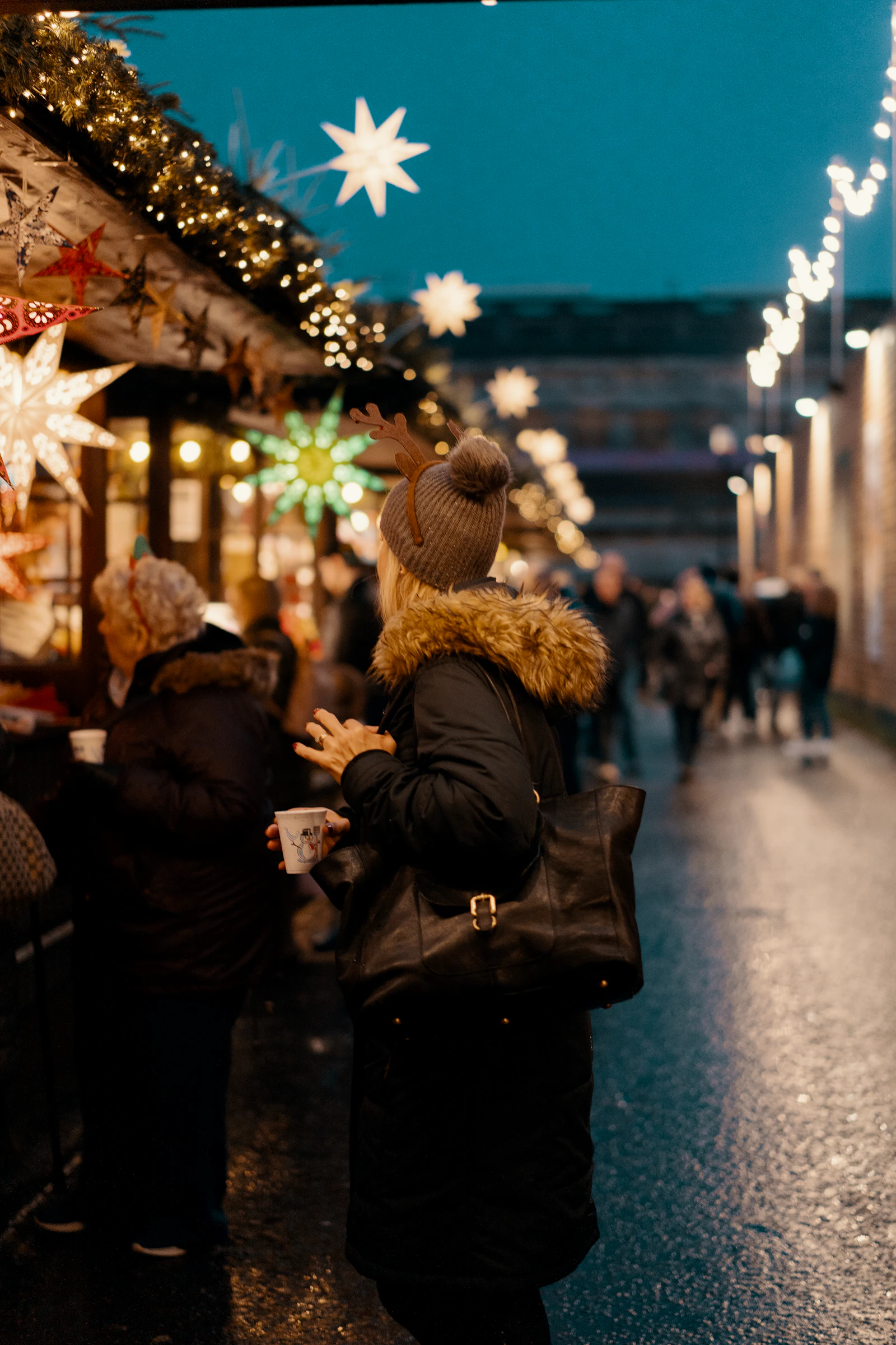 Woman in a winter coat holding a paper cup of hot drink in front of softly blurred holiday market lights
