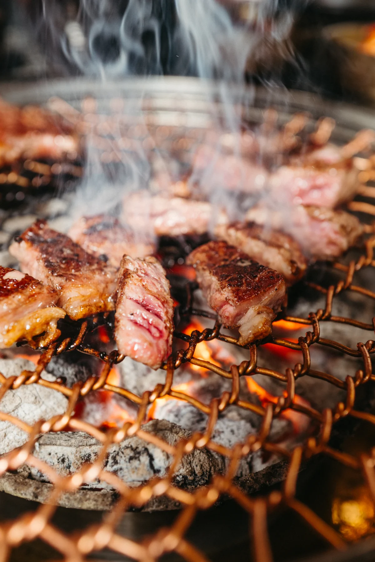 Thick cuts of beef and pork searing over a charcoal grill with smoke rising