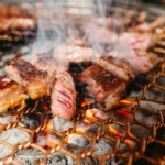 Thick cuts of beef and pork searing over a charcoal grill with smoke rising