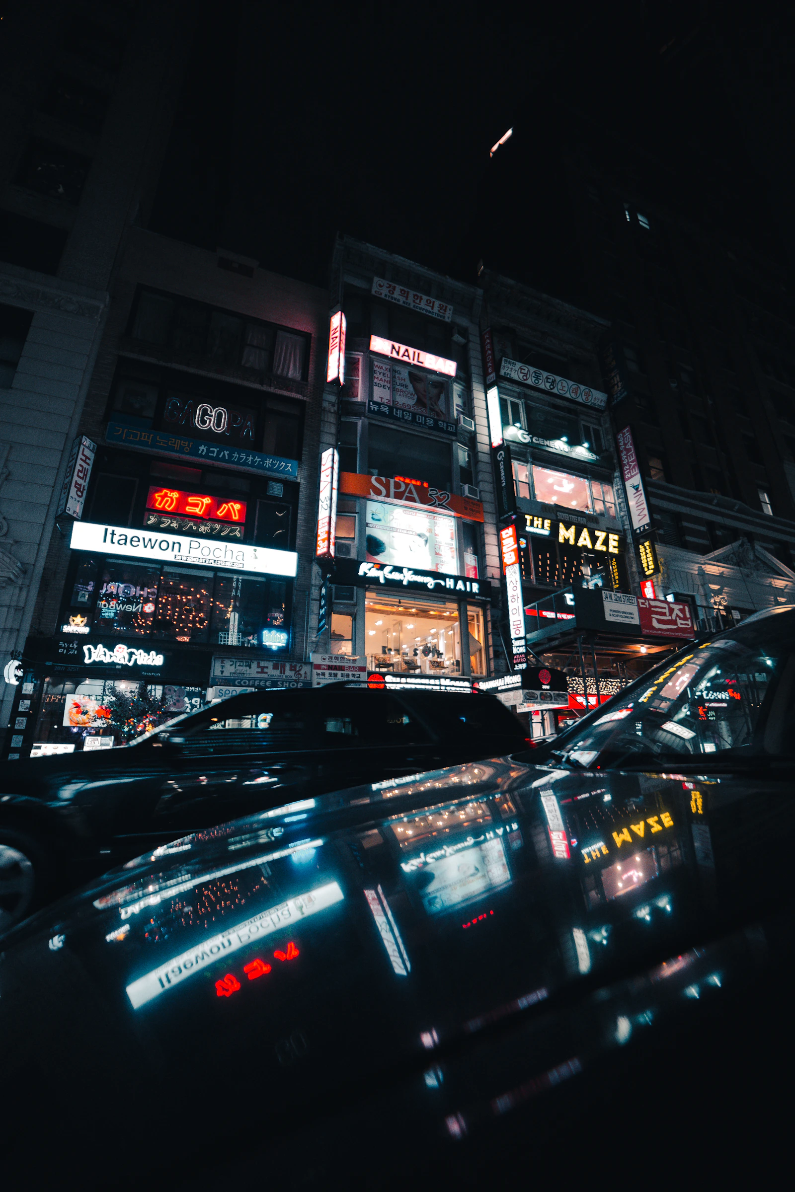 Neon Korean and English signs stacked above a Koreatown street at night with cars in the foreground
