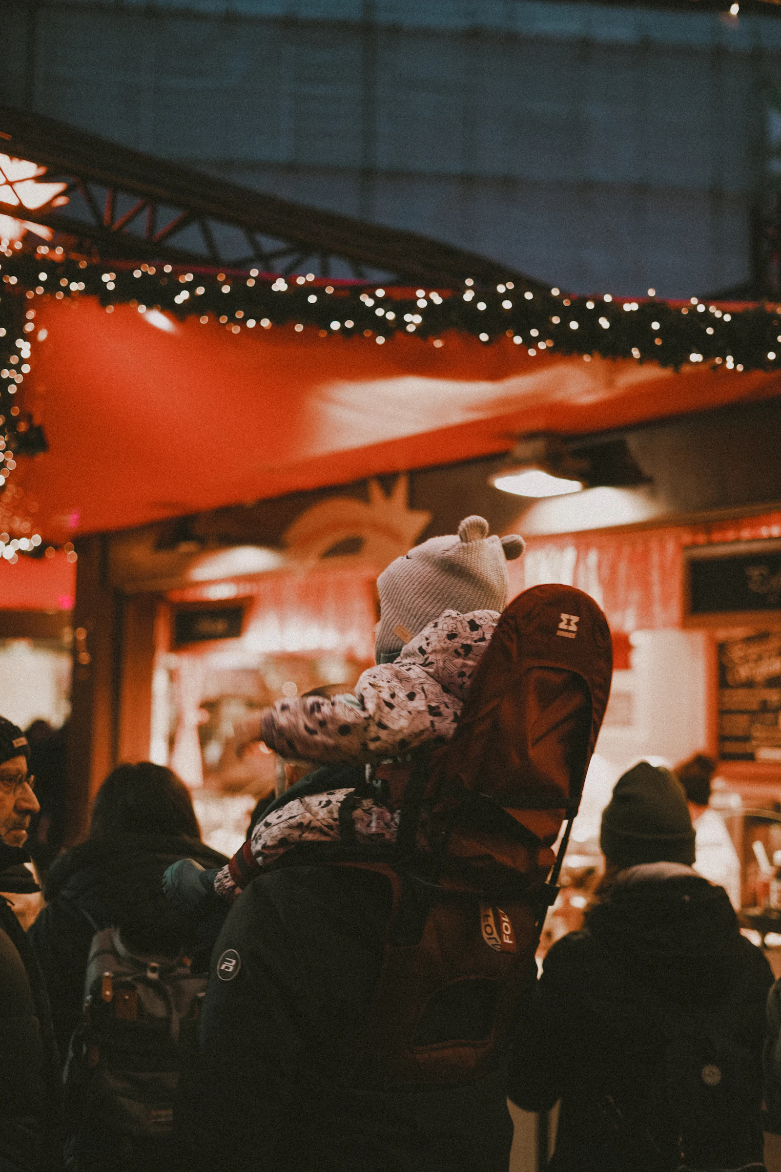A parent carrying a toddler in a baby carrier at a lit holiday market stall