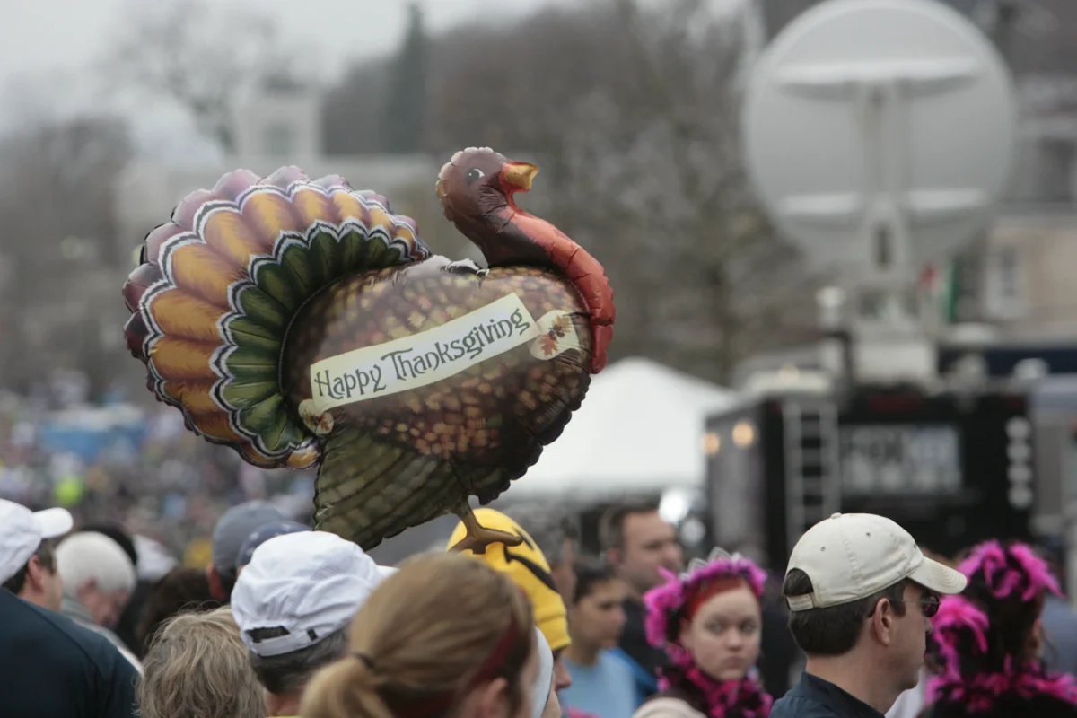 A turkey-shaped parade balloon reading Happy Thanksgiving floats above a crowd on a city street