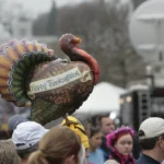 A turkey-shaped parade balloon reading Happy Thanksgiving floats above a crowd on a city street