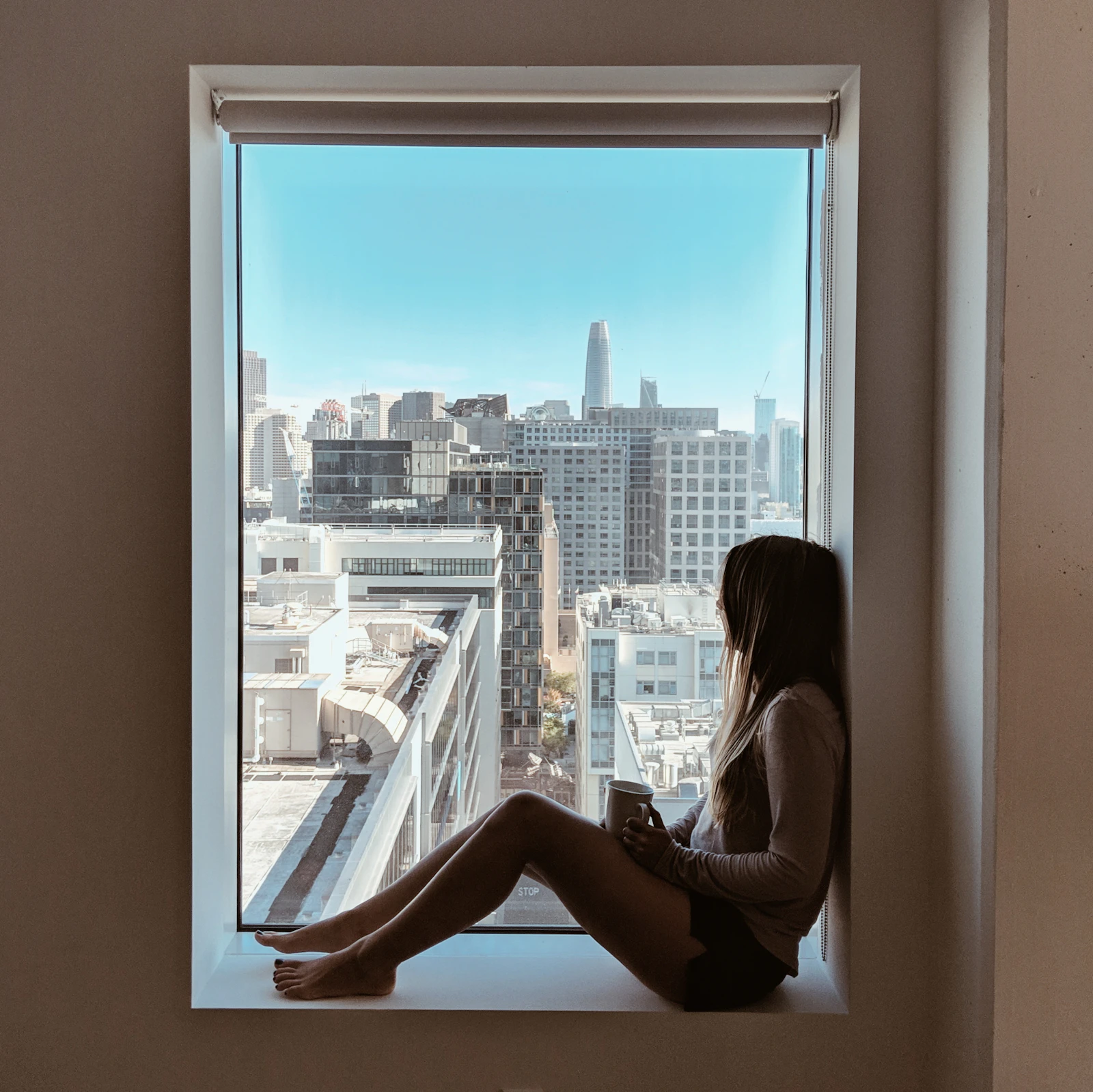 A woman sitting in a hotel room window ledge with a cup, looking out over a city skyline in daylight