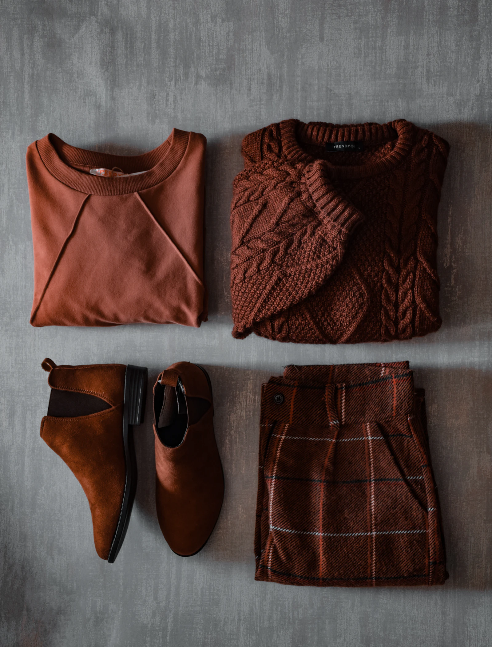 Folded rust-colored sweaters, brown suede boots and plaid trousers laid flat on a gray surface