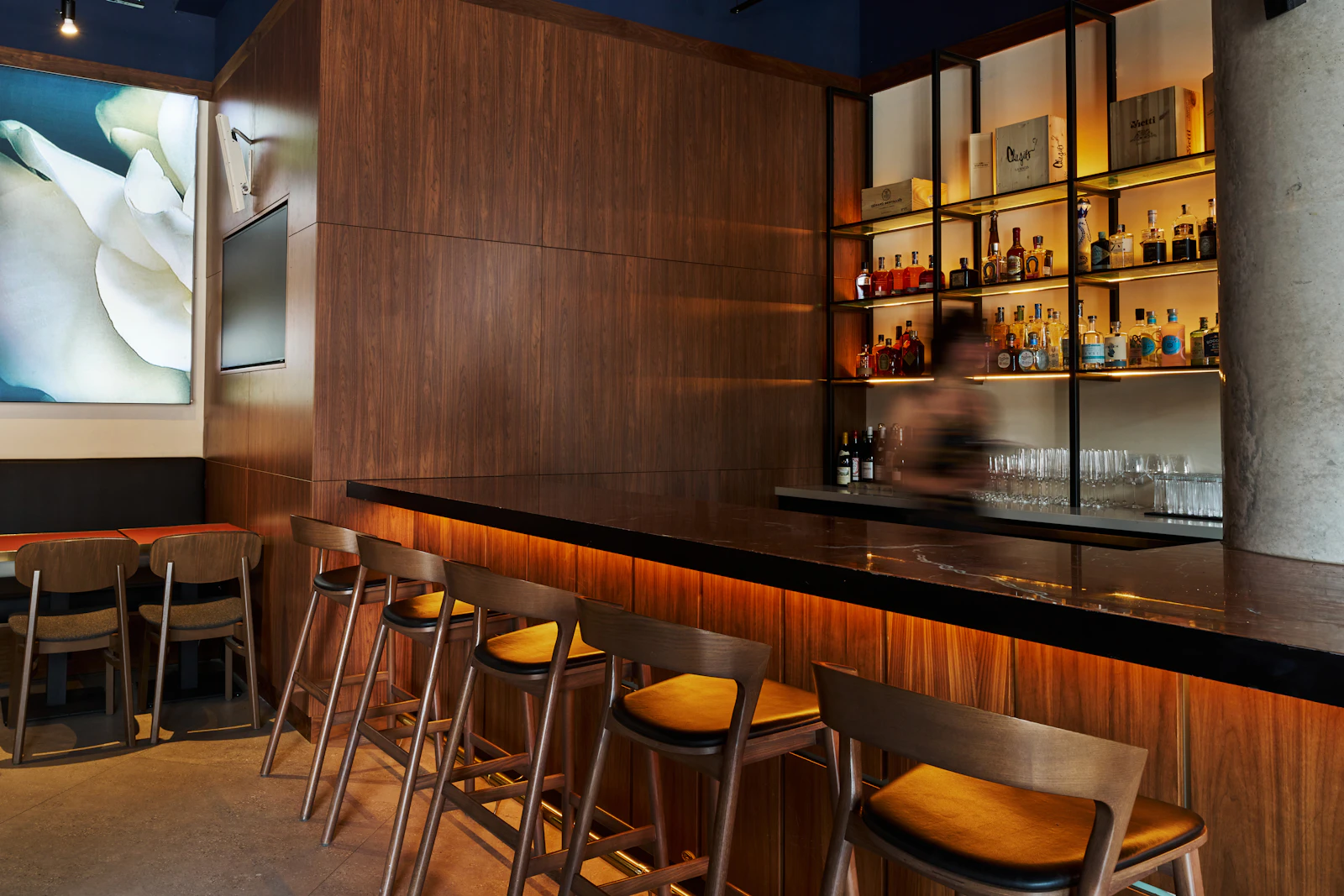 An empty wooden bar counter with a row of stools and a lit shelf of bottles