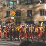 Performers in red and gold costumes march behind gold balloon letters at a Thanksgiving parade on a Manhattan street