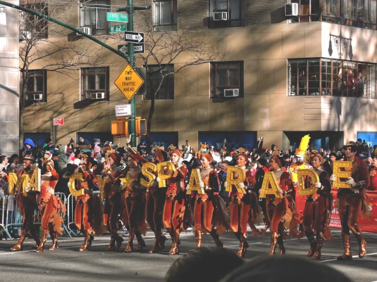Performers in red and gold costumes march behind gold balloon letters at a Thanksgiving parade on a Manhattan street